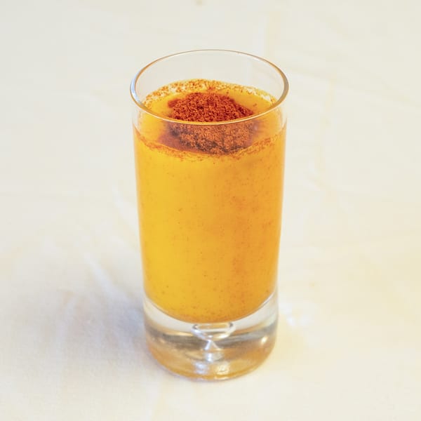 Yellow-orange drink in a tall glass, topped with reddish-brown powder, on a light background.