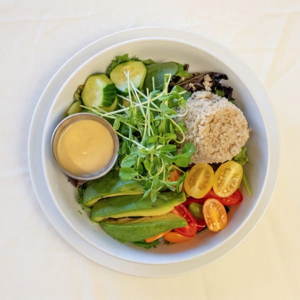 Healthy quinoa bowl with avocado, cherry tomatoes, cucumber, mixed greens, and a side of dressing.