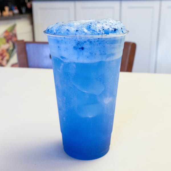 Vibrant blue foamy drink with ice in a plastic cup on a white table.