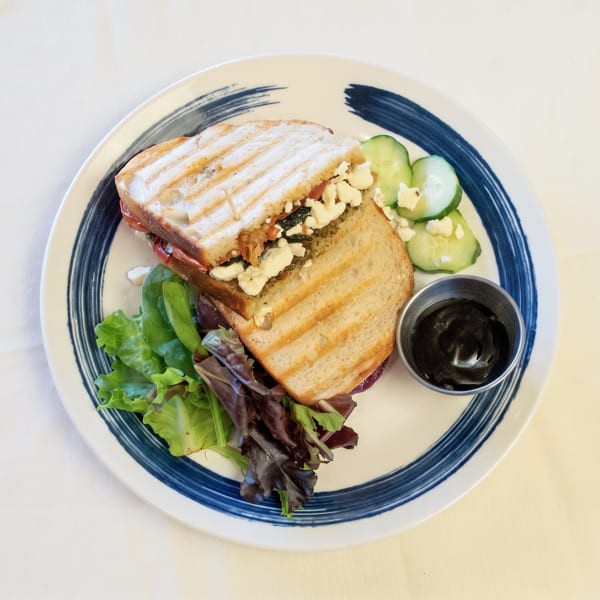 Grilled sandwich with feta, olives, mixed greens, cucumbers, and dark sauce on a blue-rimmed plate.