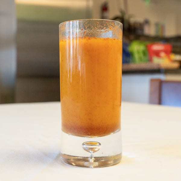 Tall glass of vibrant orange-red drink with sediment on a white tablecloth.